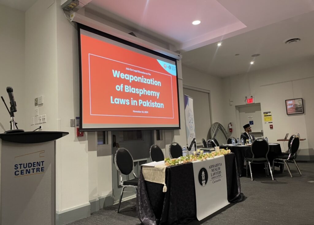 A conference room with a large projection screen displaying the title "Weaponization of Blasphemy Laws in Pakistan" in bold white text on a red background. The event is hosted by the Ahmadiyya Muslim Lawyers Association Canada.