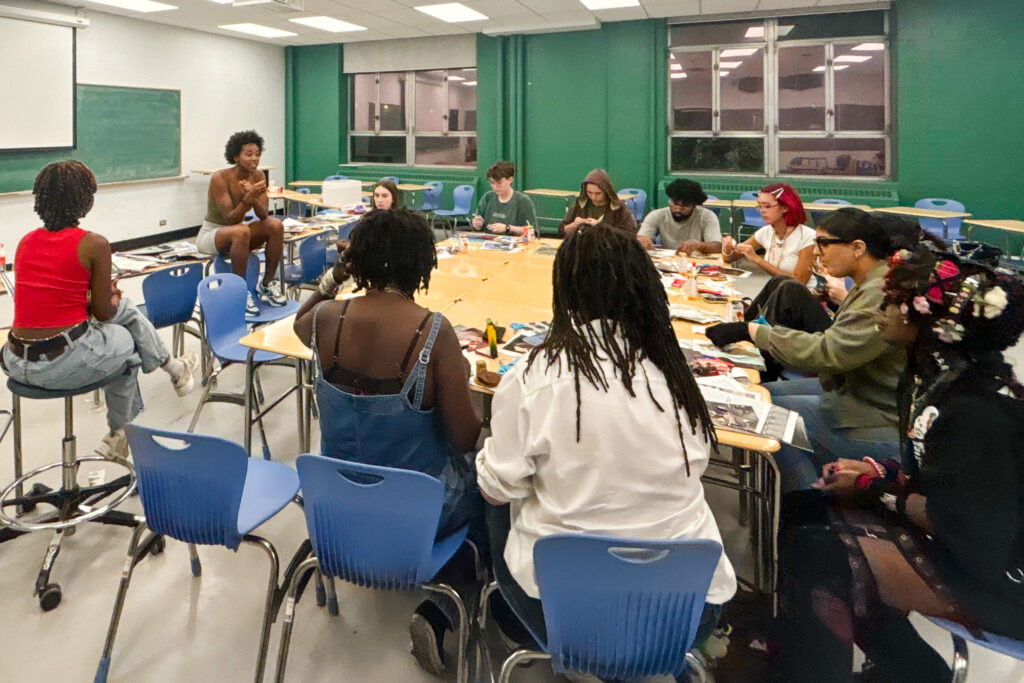 A group of students sitting around conjoined tables with scattered magazines and art supplies. One person sits atop a desk and speaks to the group with hand gestures