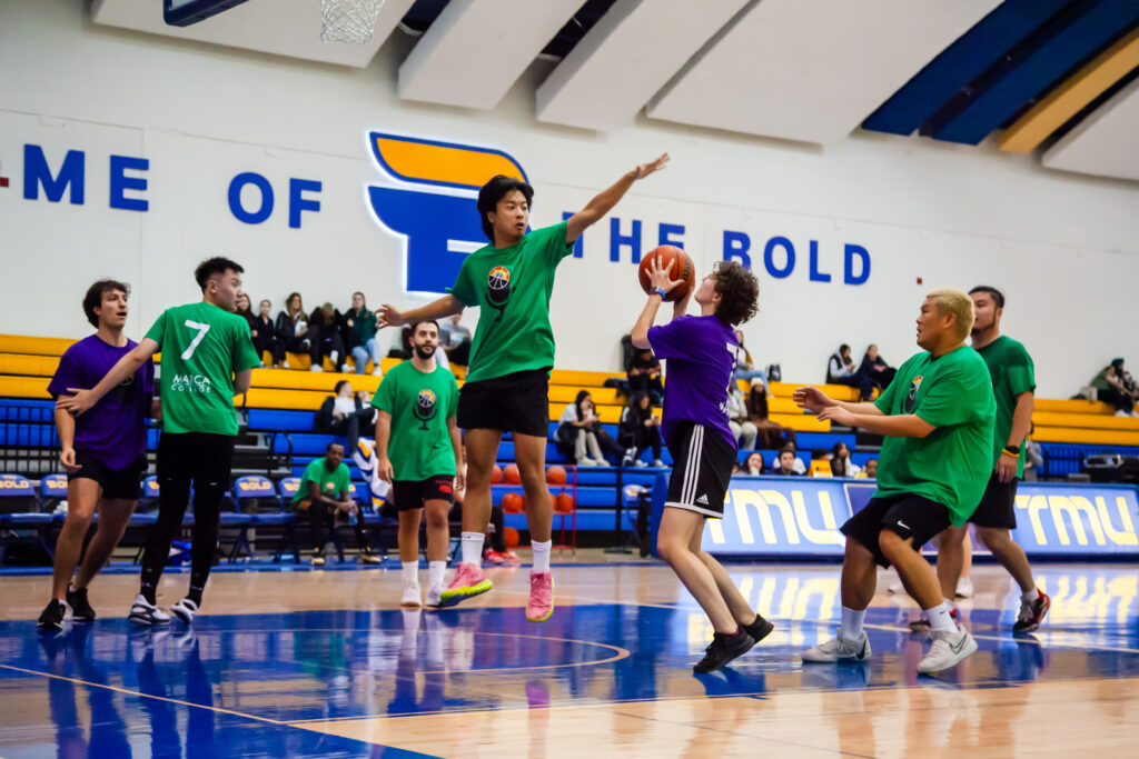 A group of basketball players on a court, some wearing green shirts and others wearing purple. A player in a purple shirt aims with a ball while another in green tries to block the shot.