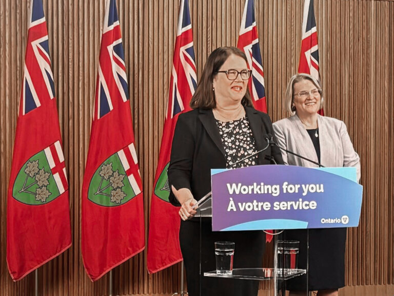 A photo of former federal health minister Dr. Jane Philpott standing at a dais, with Health Minister and Deputy Premier Sylvia Jones behind framed by a set of flags.