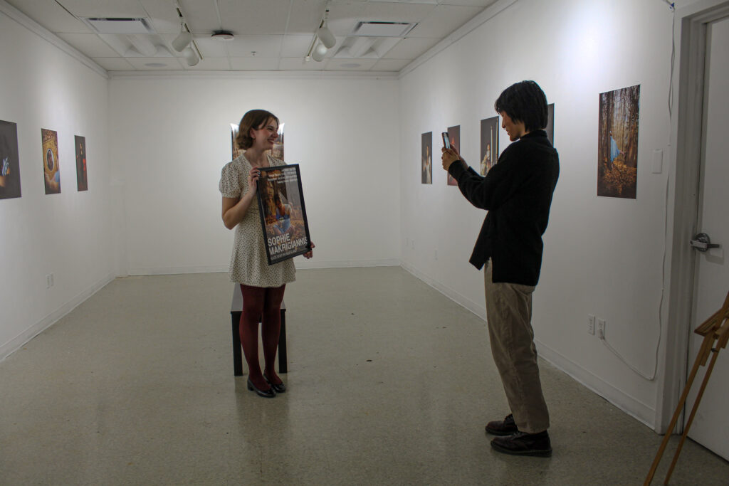 A man takes a photo on his phone of a young woman posing with a framed poster.