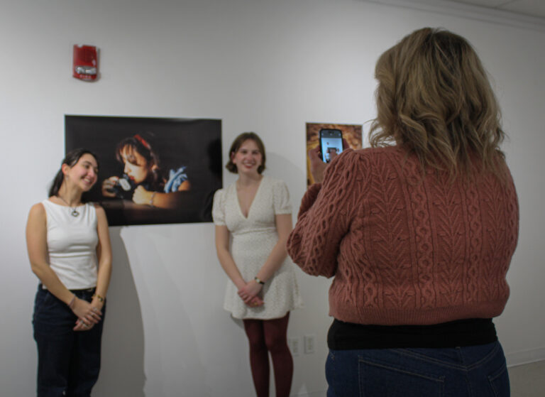 A blonde woman in a pink sweater takes a photo on her phone of two young women standing in front of a photo on the wall.