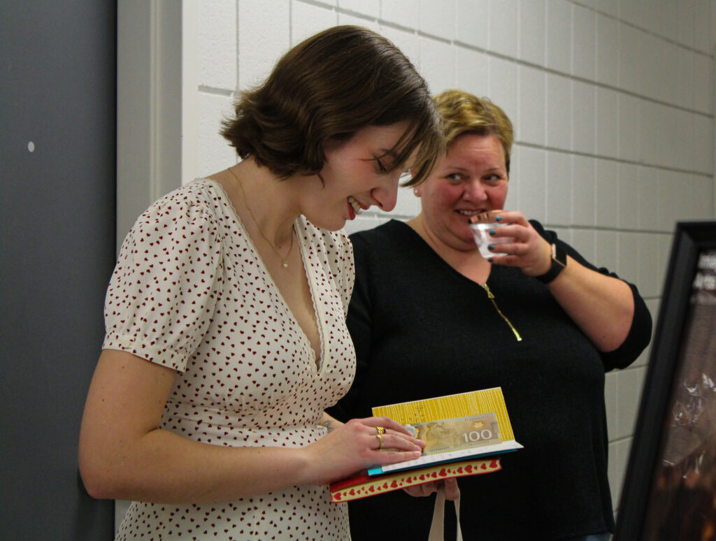 A woman with short hair drinking from a cup looking at a young woman reading a card.