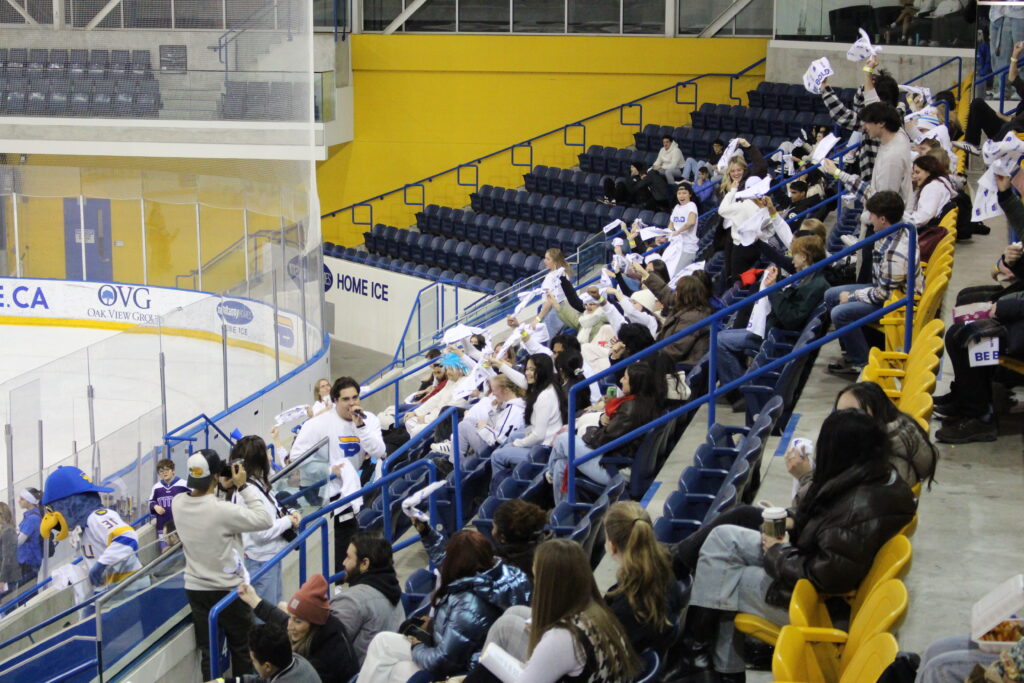 Fans in stand from their seats at an ice hockey rink, mostly wearing white tops and waving around white towels looking in the direction of a man with a microphone who is holding a white crewneck.