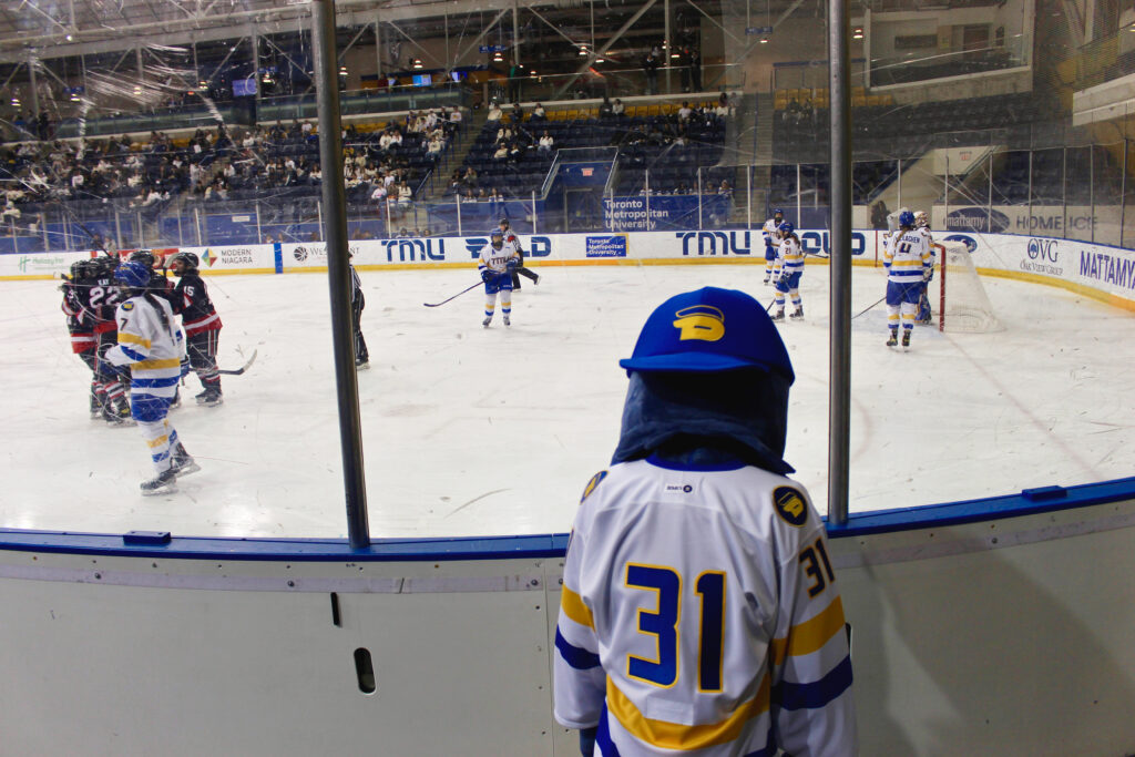 TMU mascot Frankie the Falcon has his head looking down outside the glass of the hockey rink at the Mattamy Athletic Centre where players in white jerseys face off players in black jerseys.