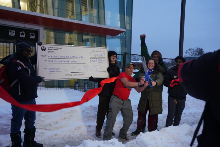 Group of people are holding a large signed cheque standing behind a building cutting red ribbon with an oversized scissors