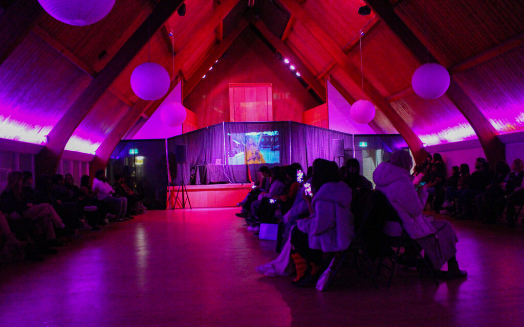 A crowd sits in rows inside a room with cathedral ceilings lit with pink and red lights