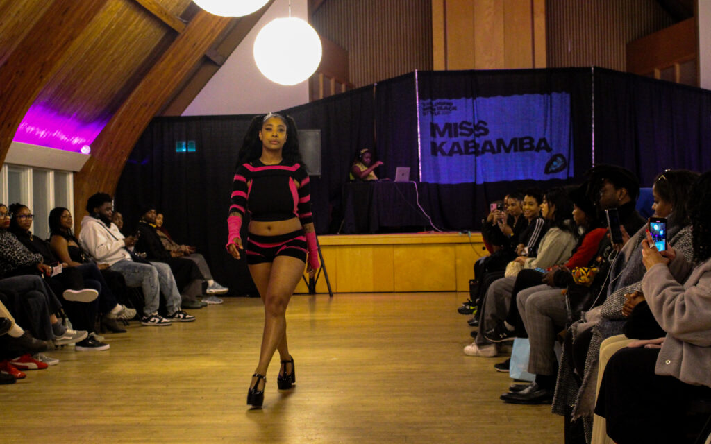 A woman with pigtails and a black and pink crop top and shorts set walk down a runway.