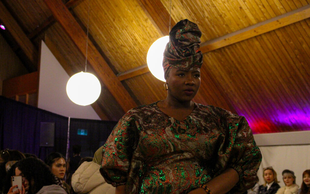 A woman in a green and gold dress and matching headwrap poses at the end of a runway