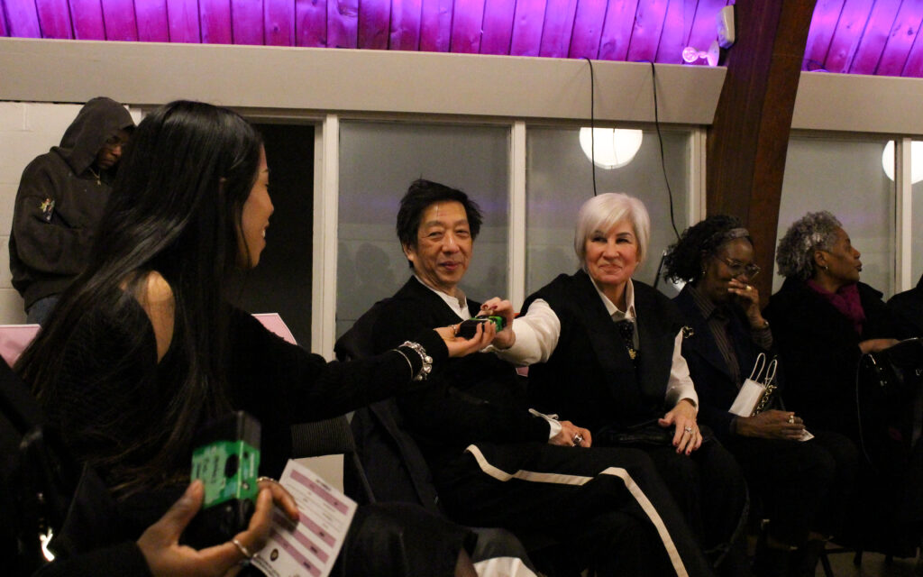 A wooman with white hair passes a green disposable camera to a woman sitting two seats down from her