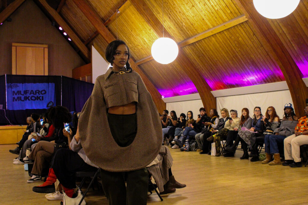 A woman wearing a cropped brown jacket with connected sleeves poses at the end of a runway