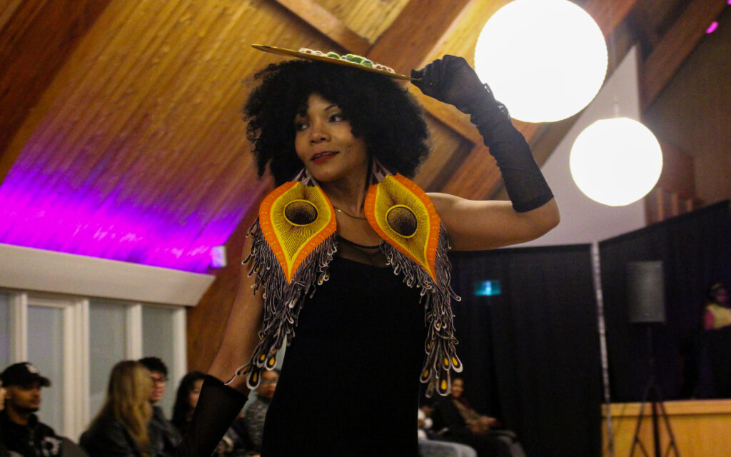 A woman wearing a black dress, gloves, hat and big orange and yellowfeather earrings poses on the runway