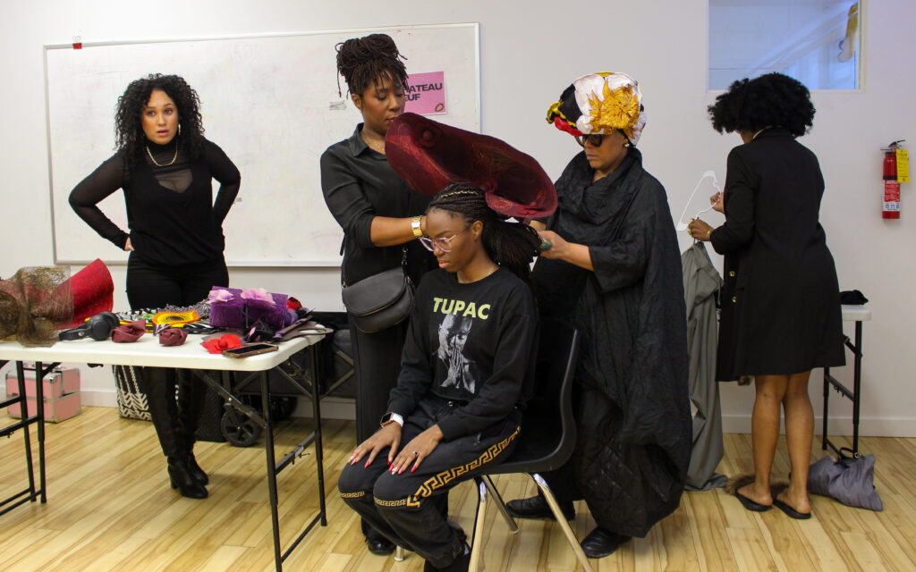 Two women help fit a headpiece onto a sitting model, while two other women stand on either side of them.