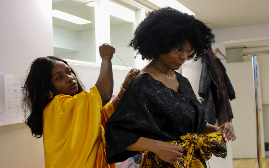 A woman in a yellow dress sews the top of the black shirt on a woman with glasses