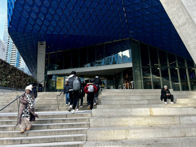 Students walk up outdoor concrete stairs leading up to a glass entrance to a geometric building with the words "Sheldon and Tracy Levy student learning centre" on the front. The students are wearing backpacks and winter attire such as coats, scarves and hats.