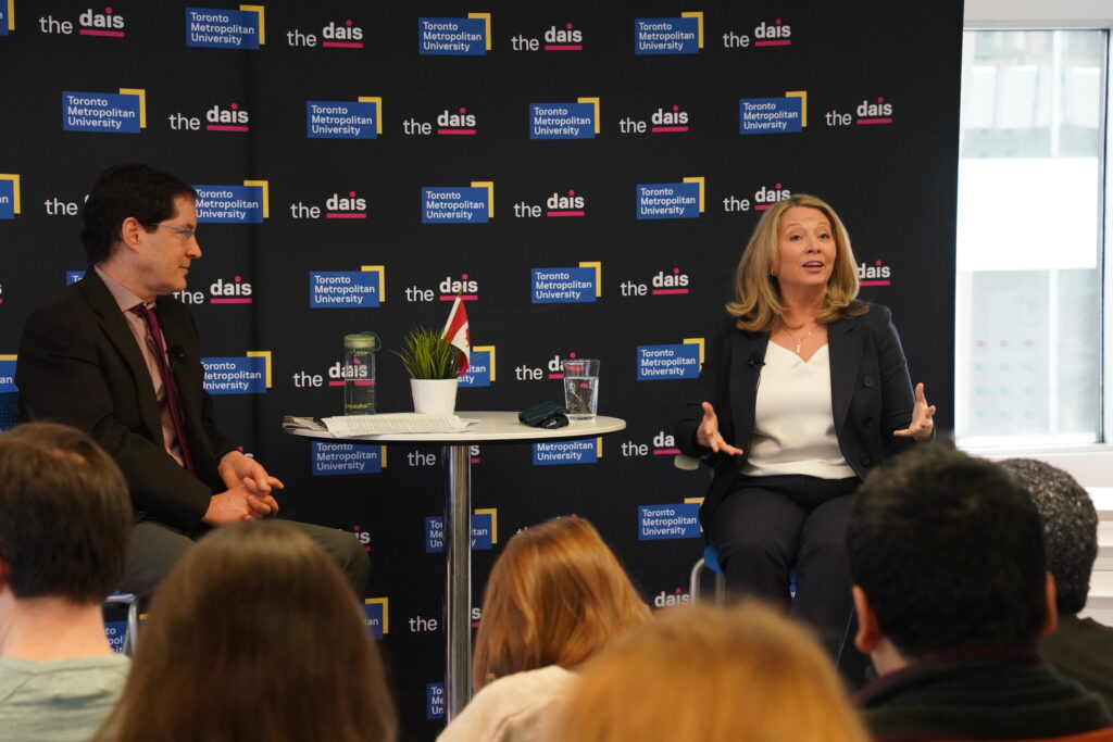 A blonde woman (Marit Stiles) sits on a high chair facing an audience of people. Next to Marit is a white high table with a glass of water, a plant with a Canadian flag and a stack of papers. To the left of the high table is a brown-haired man (Martin Regg Cohn) who wears a suit and is looking at Marit. Behind the man and the woman is a black drop sheet with the Toronto Metropolitan University blue and yellow logo and The Dais logo in white with magenta accents.