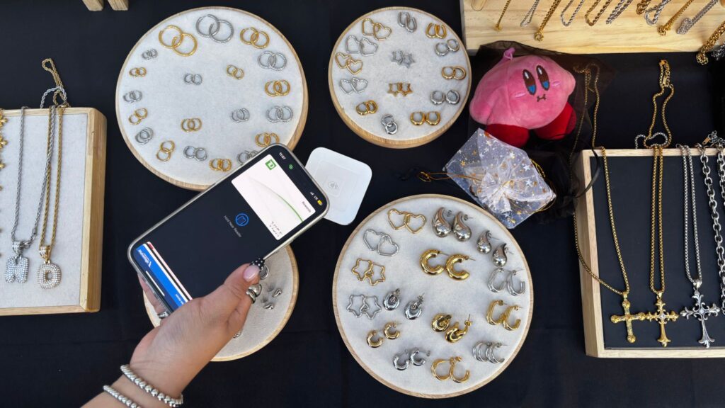 A person pays with their phone at a jewelry stall displaying gold and silver earrings, necklaces, and a small pink plush.