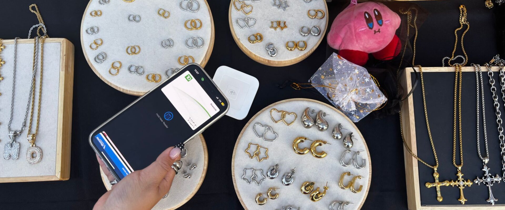 A person pays with their phone at a jewelry stall displaying gold and silver earrings, necklaces, and a small pink plush.