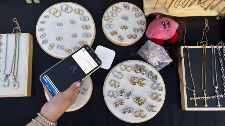 A person pays with their phone at a jewelry stall displaying gold and silver earrings, necklaces, and a small pink plush.