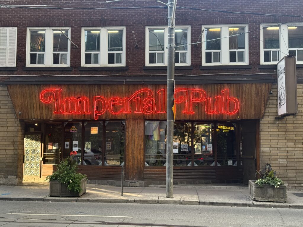 A photo of the external front entrance of the Imperial Pub featuring its iconic red neon sign above large street-level windows.