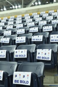 A photo of multiple rows of empty seats at the Mattamy Athletic Centre covered in white towels with the TMU Bold logo and “Be Bold” written across them.