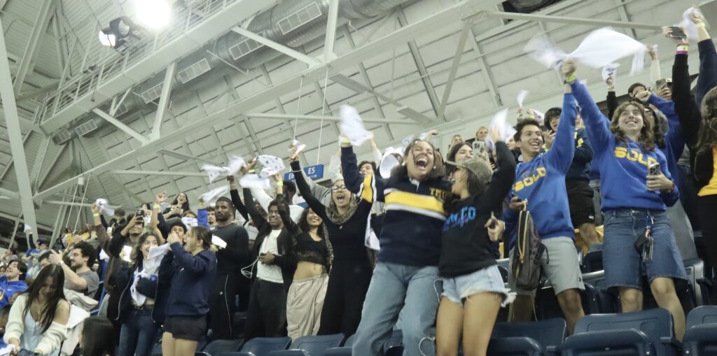 A photo of people wearing blue and white, standing up, cheering, and waving their TMU Bold towels.