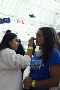A photo of two girls standing across from each other, and one is painting the other’s face blue with a Q-Tip.
