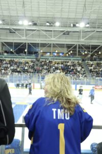 A photo of a blonde woman wearing a TMU hockey jersey leaning on a glass wall in front of the ice in a hockey arena.