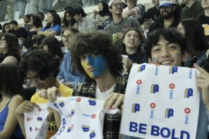 A photo of three men sitting together holding up TMU bold white towels with their faces painted in yellow and blue.