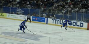 A photo of two hockey players wearing blue jerseys skating on the ice with a hockey puck.