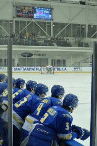 A photo of seven hockey players wearing blue jerseys on the bench leaning on the wall, watching the hockey game.