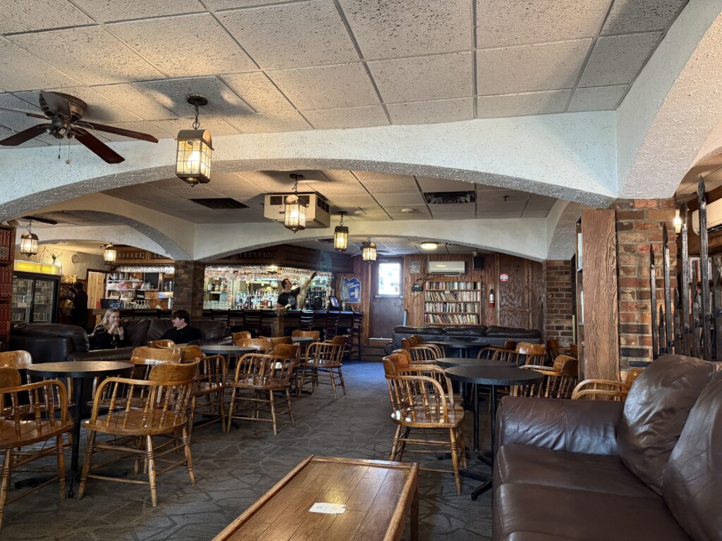 The second floor of the Imperial Pub showing wooden chairs, couches, bookshelves, and a bar with staff serving drinks in the distance.