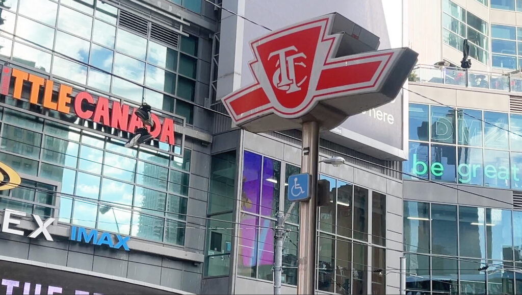 A photo of a Toronto Transit Commission (TTC) subway entrance sign with downtown Toronto buildings in the background.