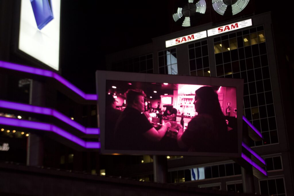 One of the screens displaying the "Disappearing Acts" video at Nuit Blanche. The screen shows footage of a same sex couple at a bar.