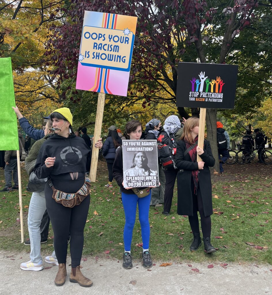 Counter-protesters hold signs reading “Oops your racism is showing,” “Stop pretending your racism is patriotism,” and “So you’re against immigration? Splendid! When do you leave?” at Queen’s Park in Toronto