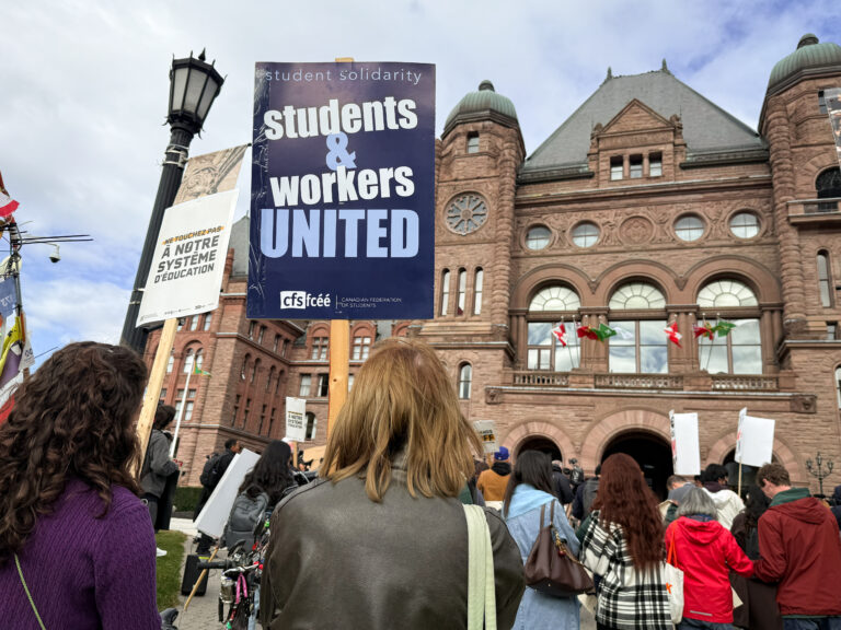 Protester holds up a sign that reads “students & workers united” in front Queens Park legislature building.
