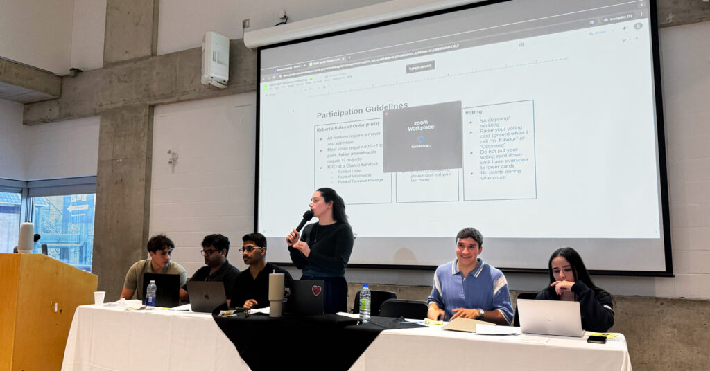 A photo of a group of five people behind a table, with one holding a microphone and speaking, against the backdrop of a projection screen.