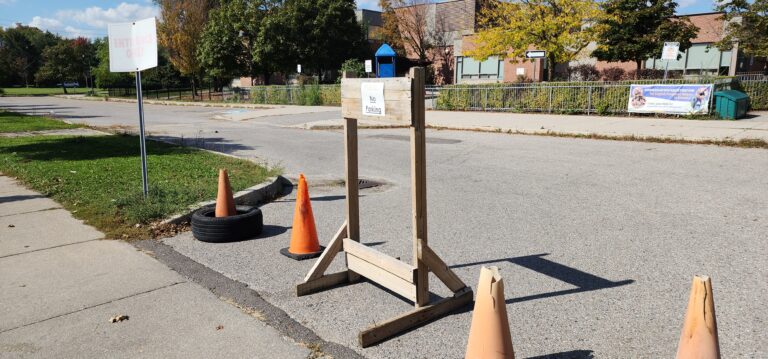 School driveway entrance blocked by orange traffic cones and a wooden sign that reads "Please No Parking" with another sign nearby marked "Entrance Only" and a school building visible in the background.