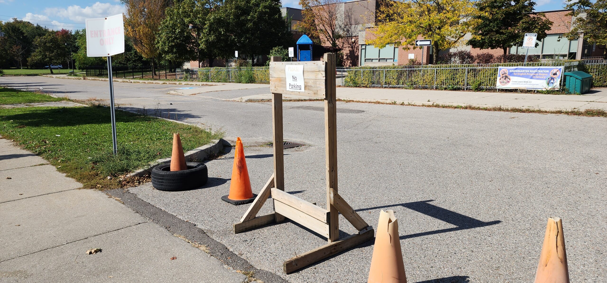 School driveway entrance blocked by orange traffic cones and a wooden sign that reads "Please No Parking" with another sign nearby marked "Entrance Only" and a school building visible in the background.