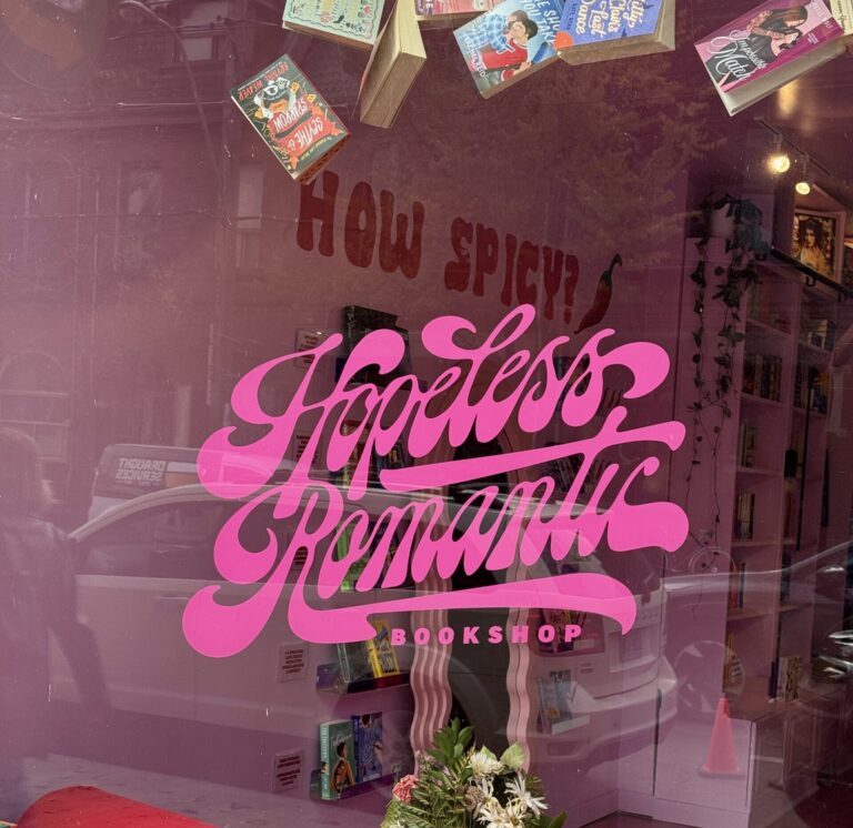 A photo of the front of the Hopeless Romantic Books bookstore. On the window pane, the name of the store is written in pink lettering. There are books hung from the ceiling.