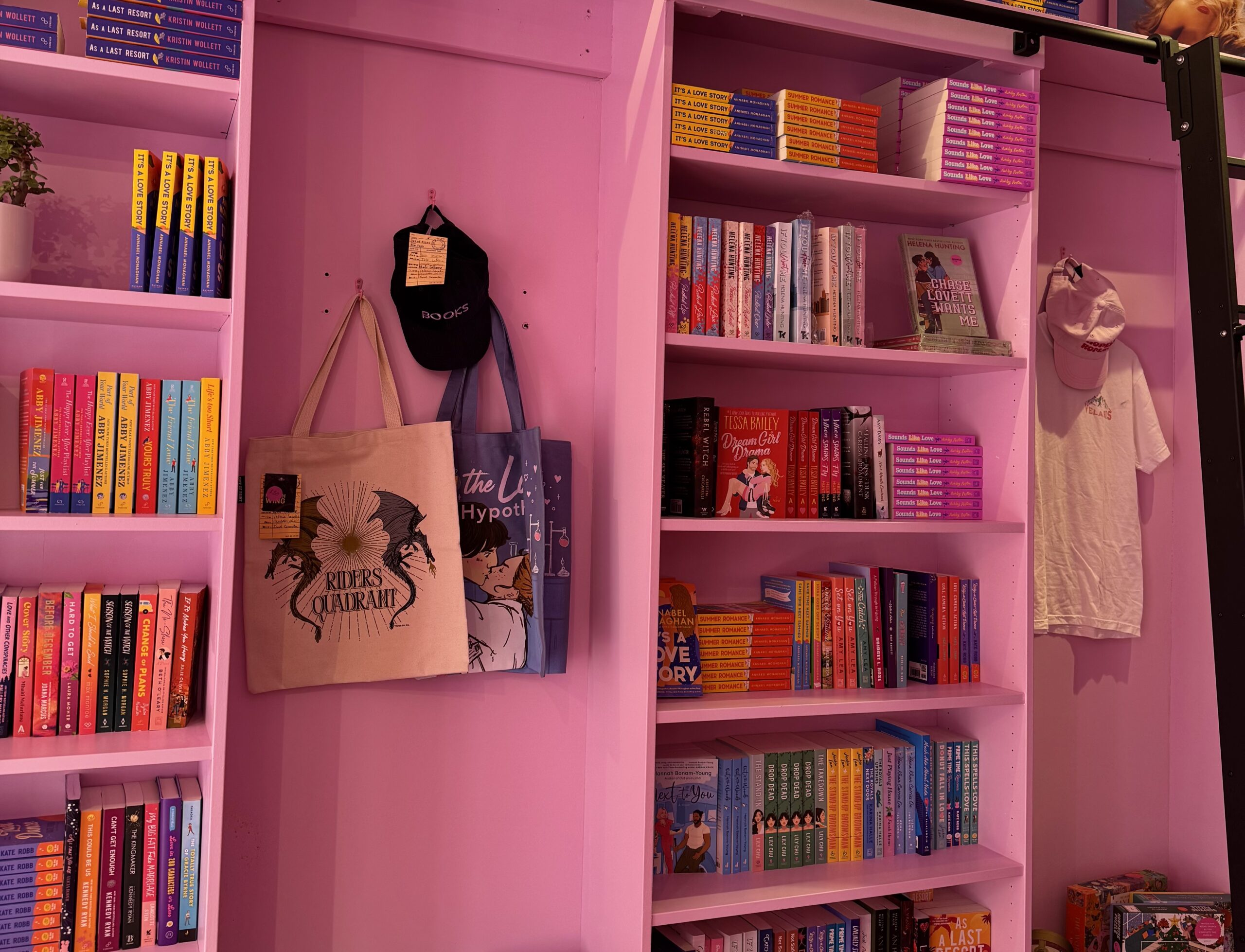 ABBAS-TIFA ROMANCE-5 The inside of Hopeless Romantic Books. There's a pink wall with pink bookshelves attached. The shelves are filled with romance books and there are totebags, hats, and a Tshirt hung on the wall.
