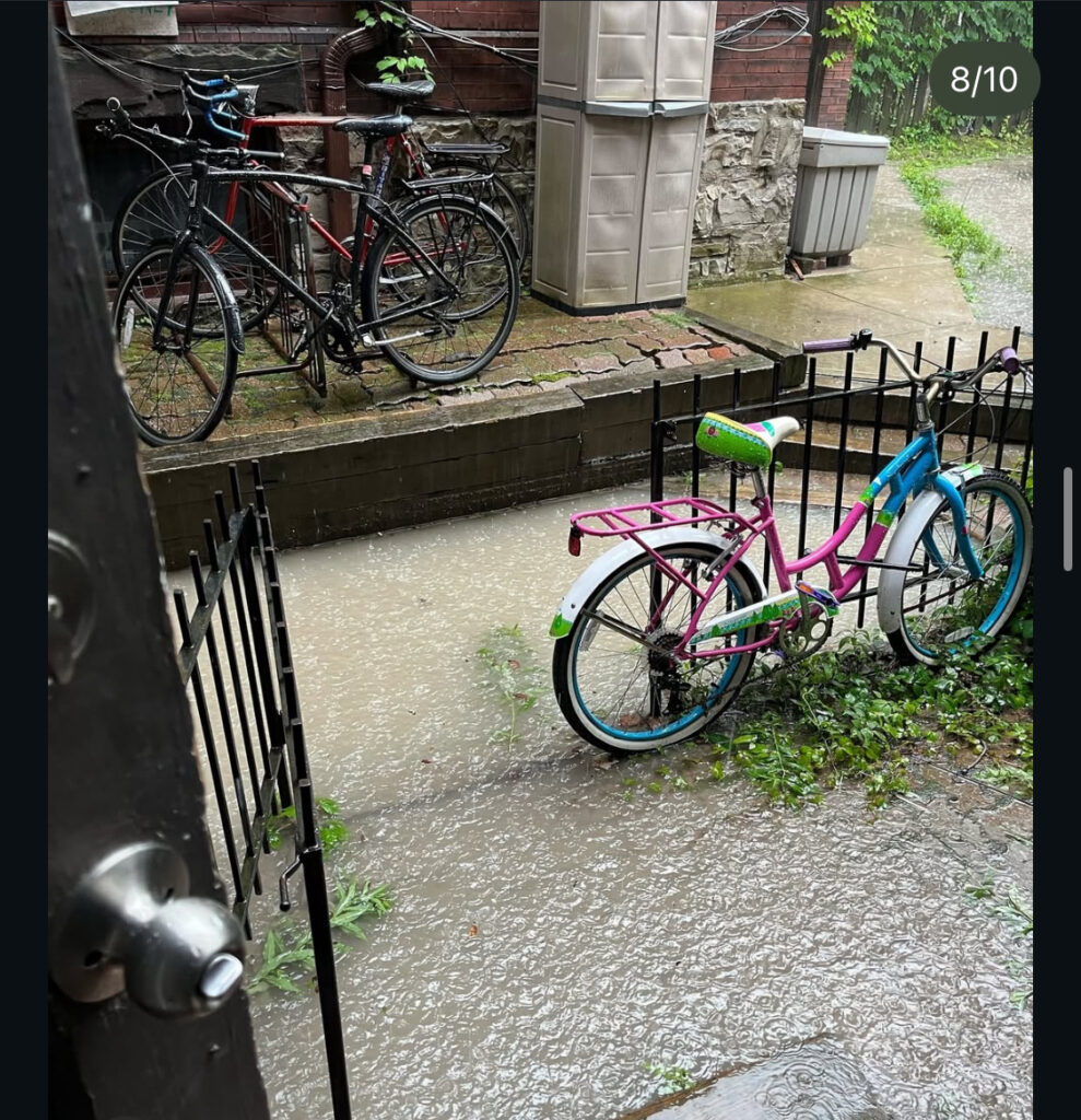 Bicycle tied to black gate during heavy rainfall, flooding the ground.