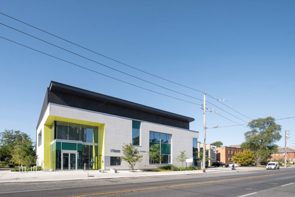 Street-level exterior view of the Mount Dennis Child Care Centre showing its large windows, bright yellow-green entrance frame and modern two-storey design.