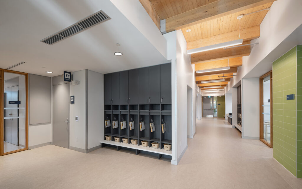 Interior hallway of the Mount Dennis Child Care Centre with exposed wood ceiling beams, built-in cubbies, classroom doors and light-coloured flooring.