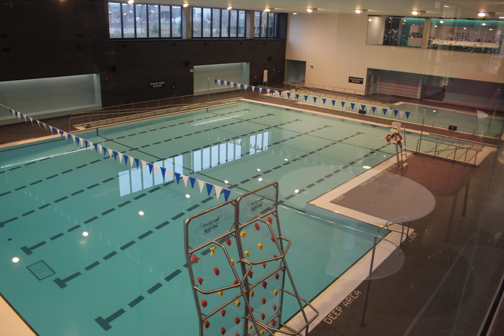 Interior view of a large swimming pool at the Rouge Valley Community Recreation Centre with lane markings, a lifeguard chair, blue-and-white flags and windows along the far wall.