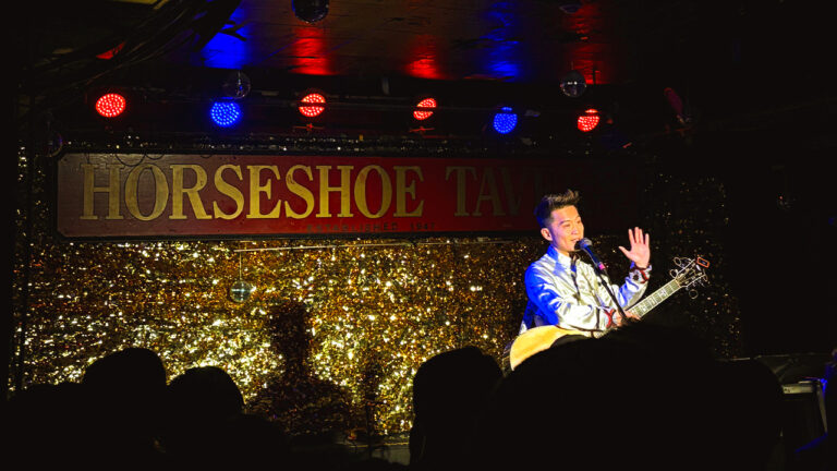 Singer/song writer Yatfung waves at audience on stage at Horseshoe Tavern in front of a sparkling gold background.