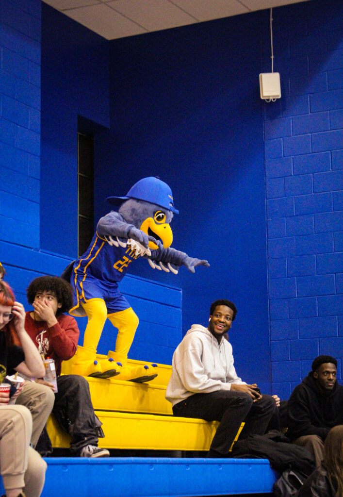 A mascot stands among spectators of a volleyball game.