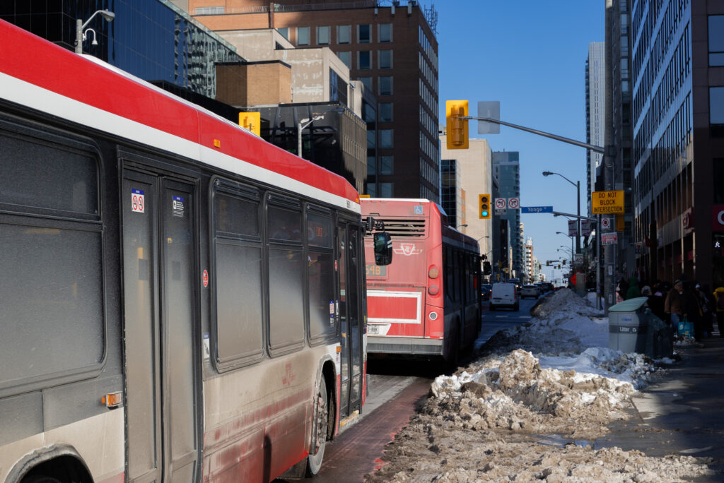 Two TTC buses covered in dirt waiting at an intersection, with a snowbank on the right side of the road.