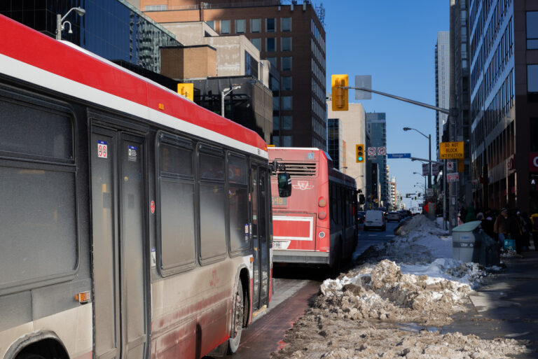 Two TTC buses covered in dirt waiting at an intersection, with a snowbank on the right side of the road.