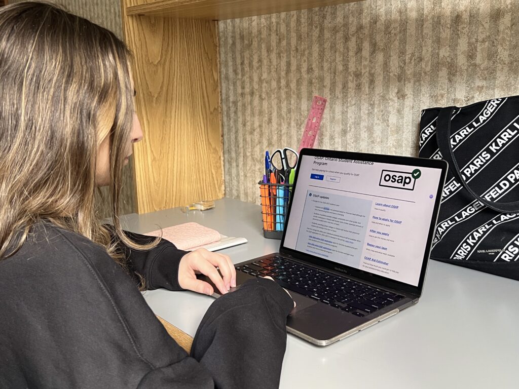 A young woman sits at a desk using a laptop displaying the OSAP website, where information about grants and loans is visible. School supplies and a notebook are beside her.